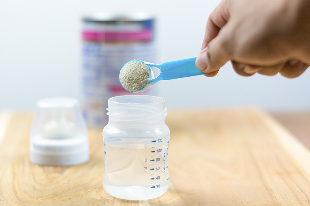 A person puts a scoop of baby formula into a baby bottle, not knowing that certain types of formula can lead to necrotizing enterocolitis.